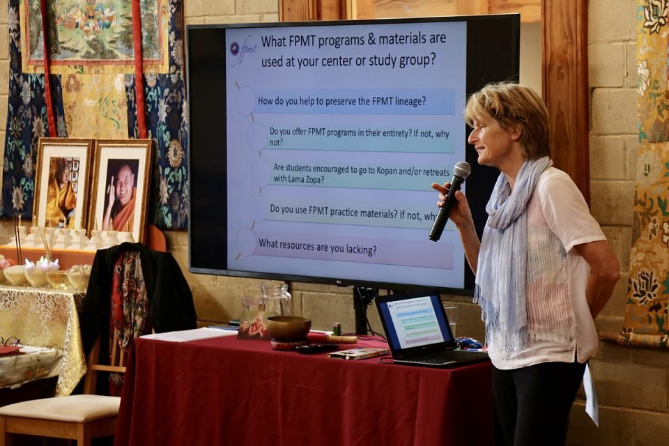 A women with a microphone stands next to a large screen in a gompa