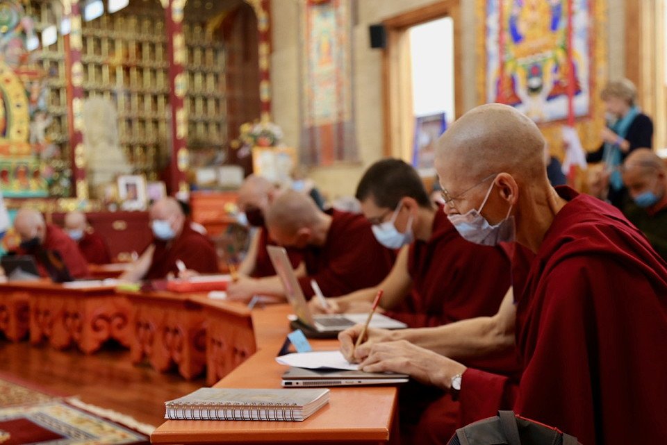 a nun wearing a mask takes notes on a puja table with other monastic and lay students in a gompa