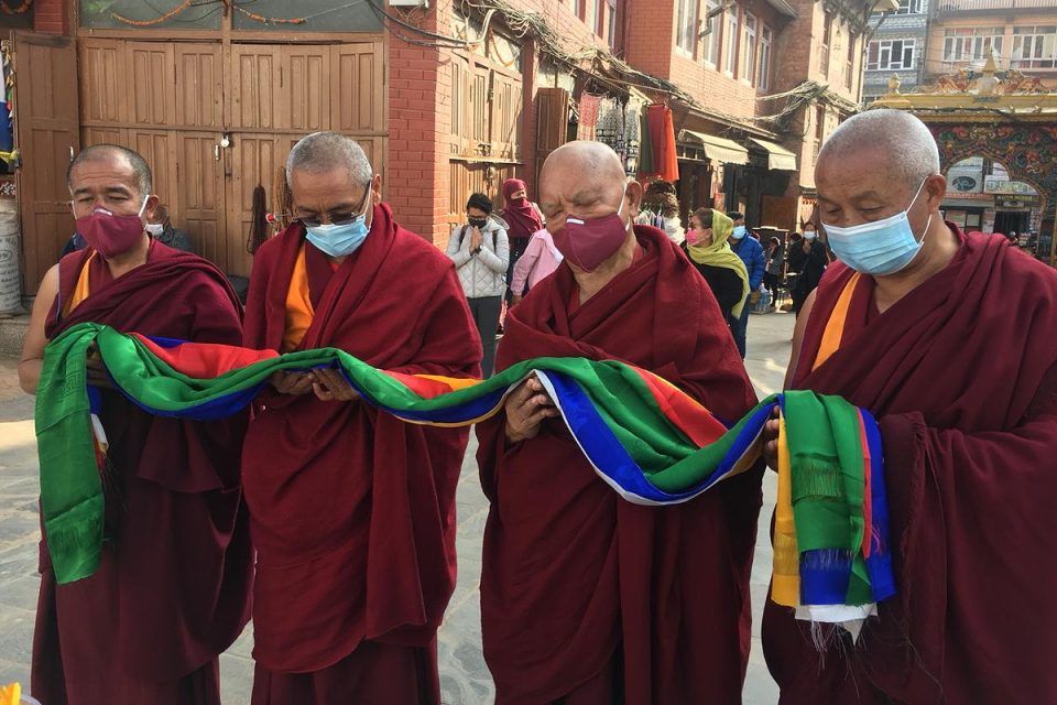 Four lamas offering five colors of khatas with brick buildings and other people in background