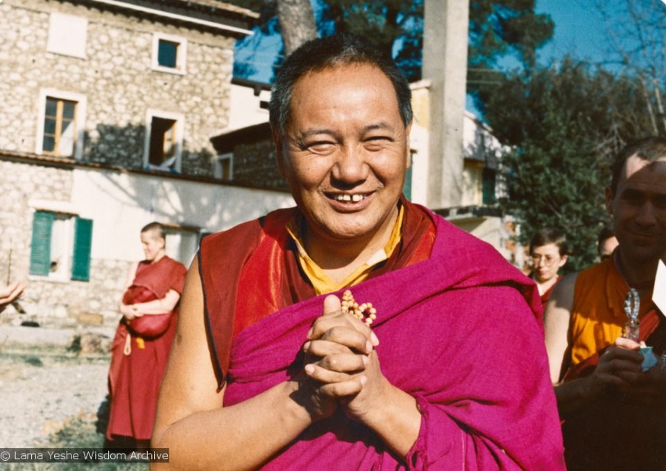 Lama Yeshe looking at the camera with hands folded around a mala with ordained sangha and an old stone building in the background