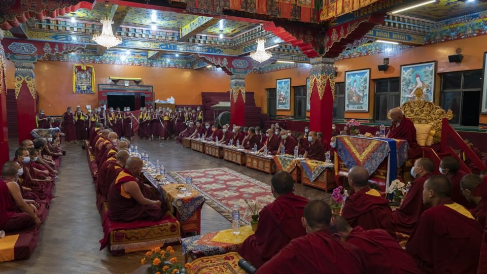monks sitting down in the debate hall with thangkhas on the wall. 