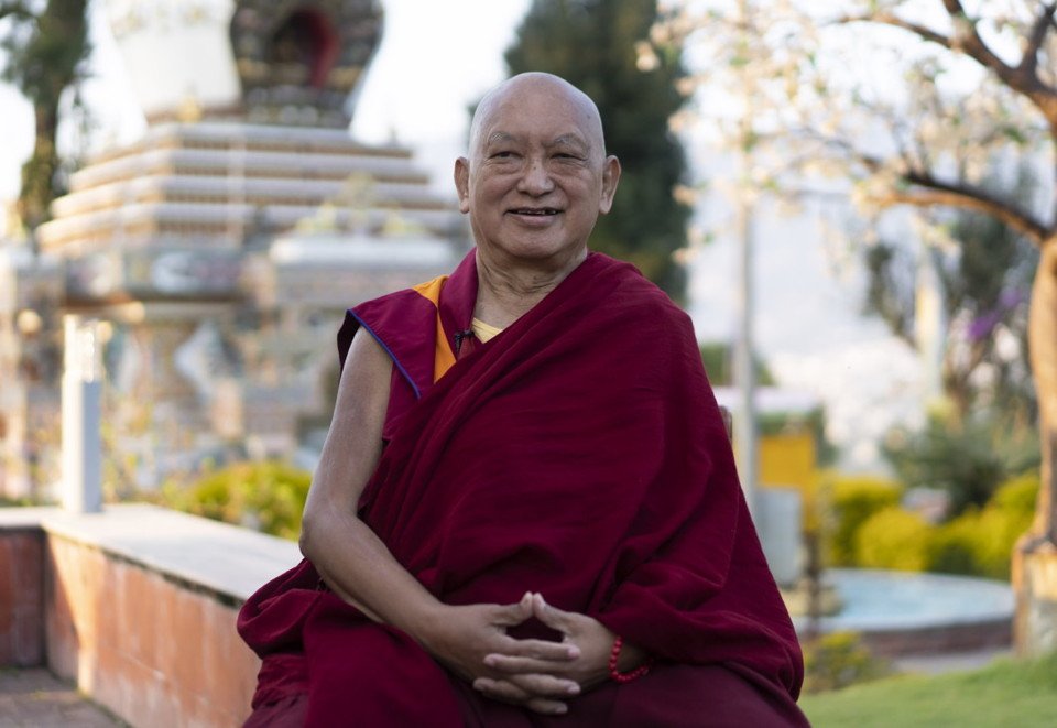 Lama Zopa Rinpoche sitting and smiling with a stupa and garden in the background
