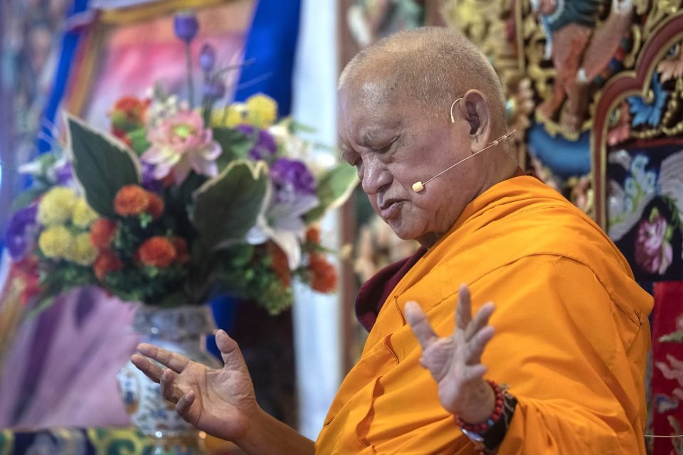 Lama Zopa Rinpoche teaching on a throne with a vase of flowers next to him