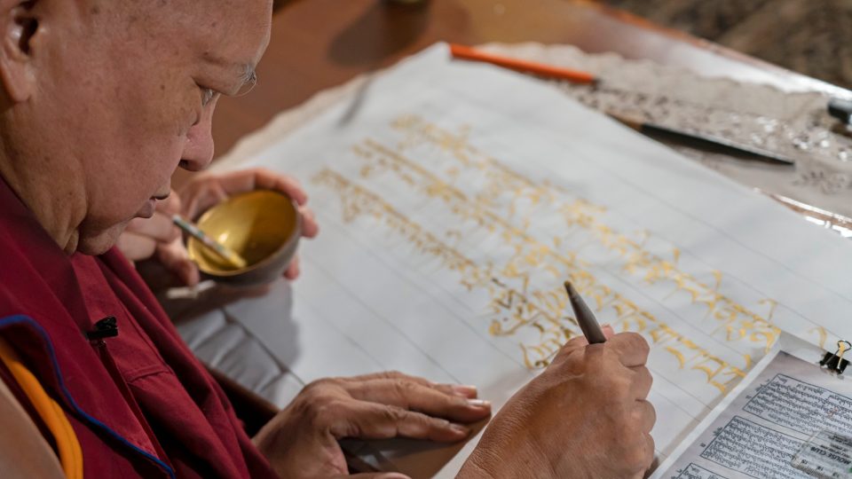 Lama Zopa Rinpoche holding a pen and writing Tibetan calligraphy on paper in gold