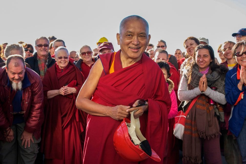 Lama Zopa Rinpoche smiling with a crowd of Sangha and students behind him. He holds a hard hat in his hand.