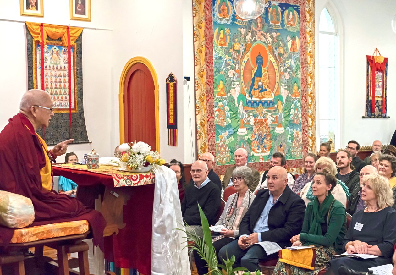 profile view of Lama Zopa Rinpoche teaching on a throne in a gompa to large audience of engaged students