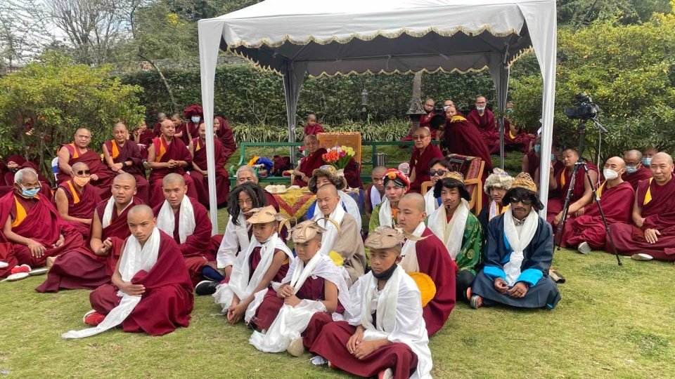 Monks in costumes with Lama Zopa Rinpoche seated under a small tent in a garden
