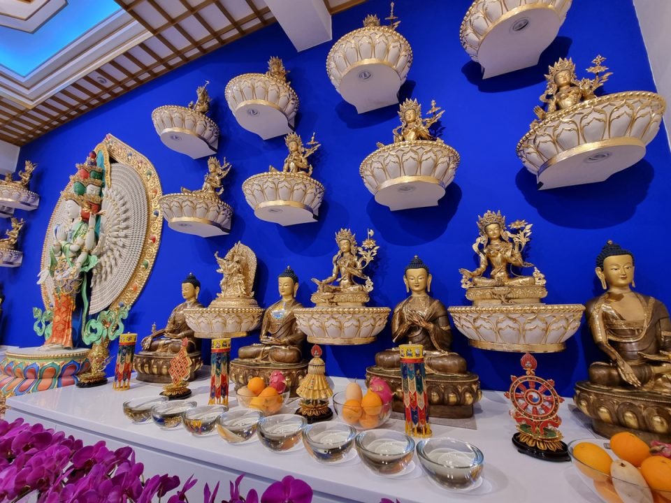 Many golden Tara statues mounted on a deep blue wall with offerings on the altar