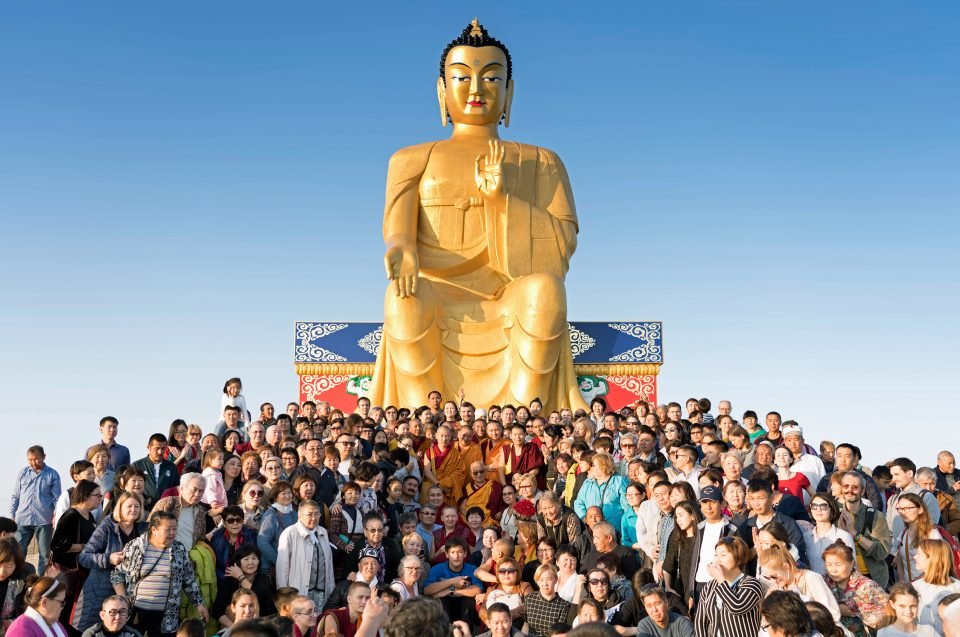 Lama Zopa Rinpoche surrounded by dozens of students in front of a large Maitreya Buddha statue under a clear blue sky
