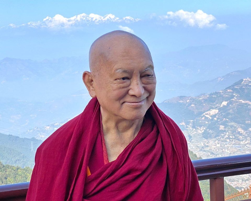 Lama Zopa Rinpoche standing on a terrace with the Himalayan foothills in the background