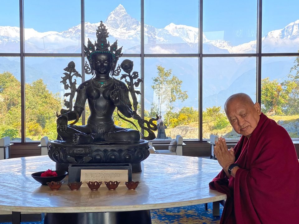 Lama Zopa Rinpoche with hands in prostration mudra next to a Tara statue with snow mountains visible through picture windows in the background
