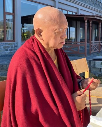 Lama Zopa Rinpoche stading outside on a terrace and holding a red mala