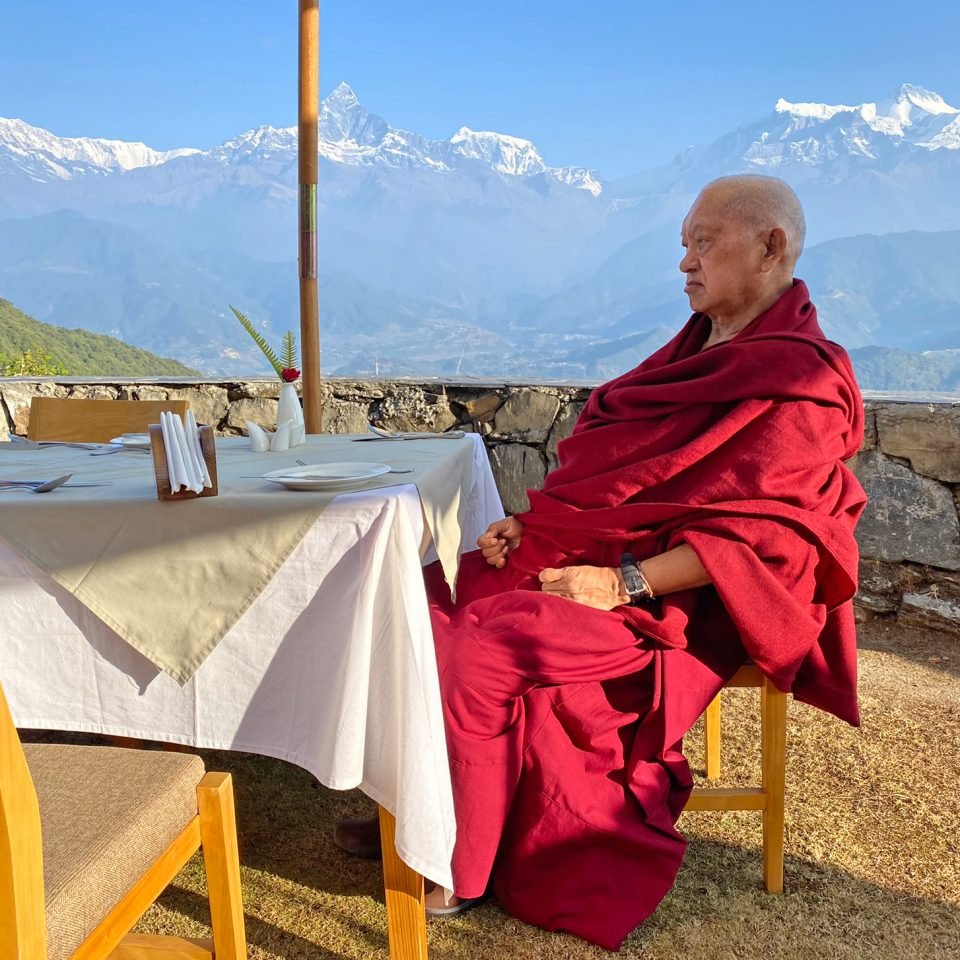 Lama Zopa Rinpoche seated at a dining table on a terrace with a view of the Himalayas