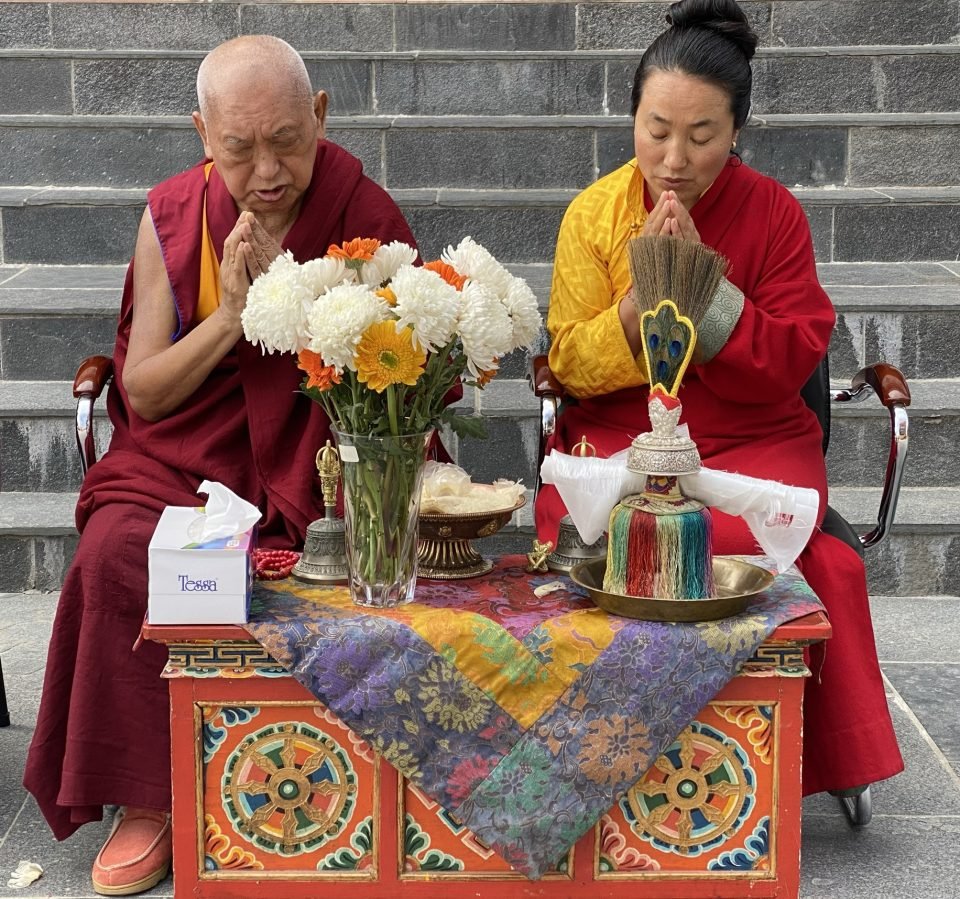 Lama Zopa Rinpoche and Khadro-la sitting at a puja table with hands in prostration mudra