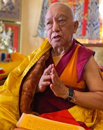 Lama Zopa Rinpoche with hands together and a text on his lap
