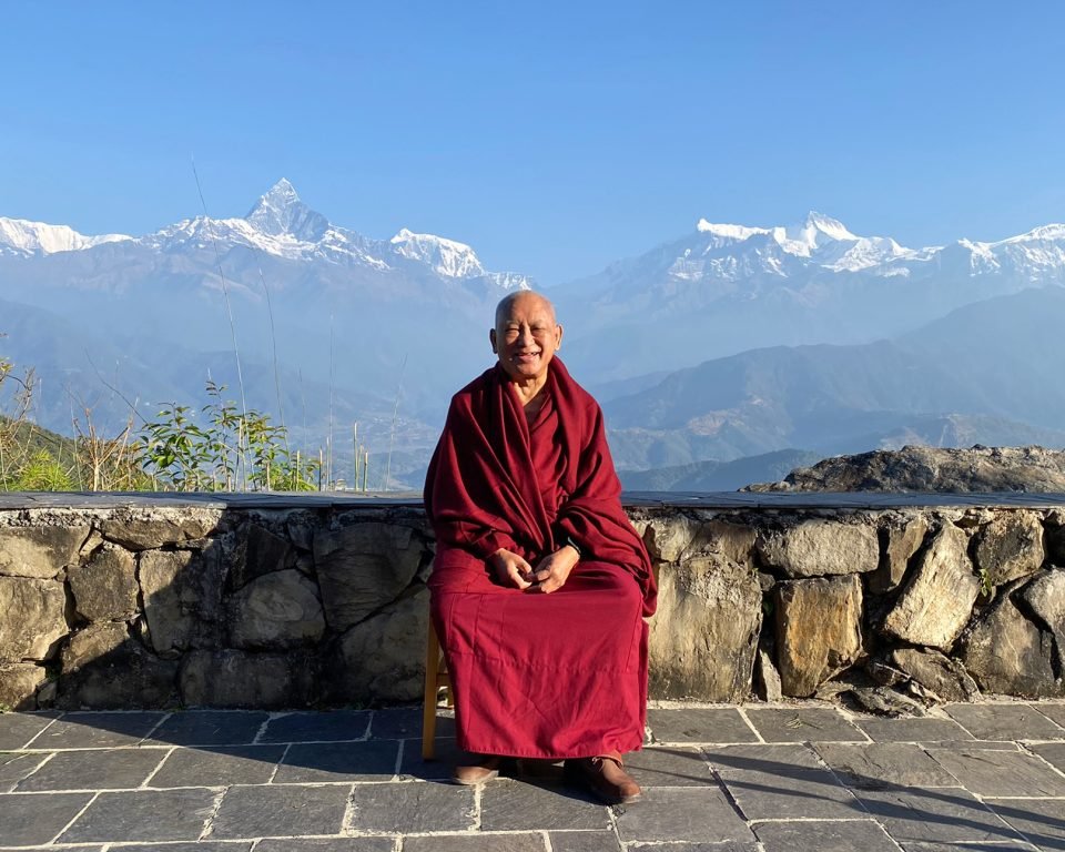 Lama Zopa Rinpoche seated on a patio with the Himalayan Mountains in the background
