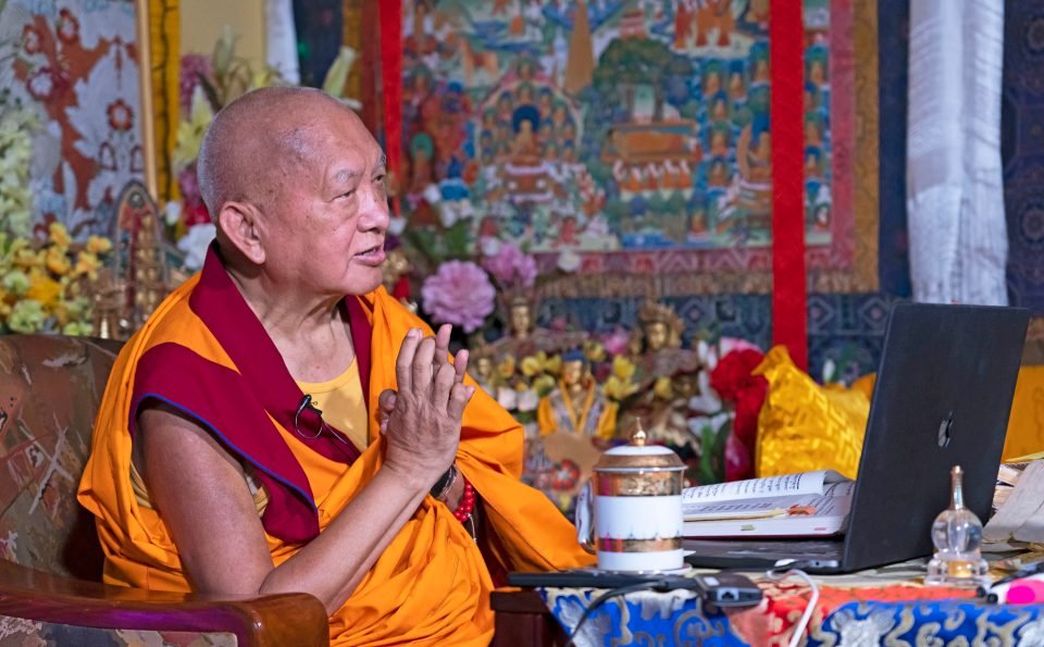 Rinpoche with hands in prostration mudra teaching in front of a laptop