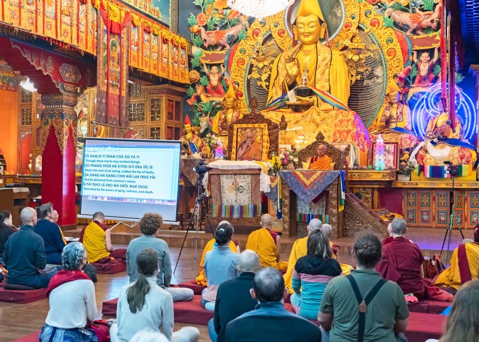 Lama Zopa Rinpoche seated on a teaching throne in front of a large Lama Tsongkhapa statue in the main gompa at Kopan Monastery