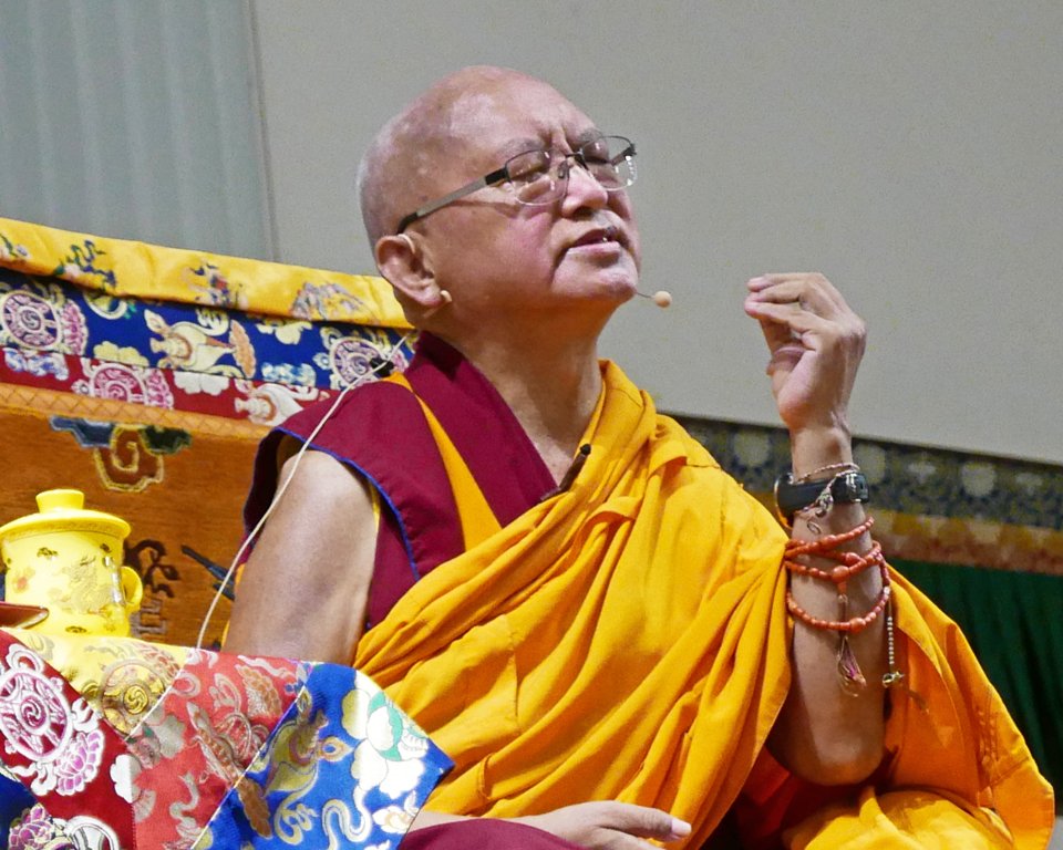 Lama Zopa Rinpoche teaching on the throne. Rinpoche's eyes are closed and he is gesturing with his hand.