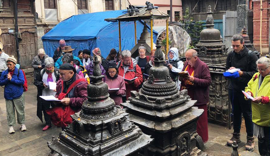 Gen Thubten Dondrub and a small group of students standing in a city square in Nepal at the Jana Bahal temple reciting from books they are holding while standing in front of a group of black colored stupas.