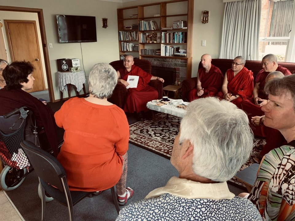 His Eminence Ling Rinpoche seated on a couch in a small room in the hospice surrounded by seated people eagerly leaning towards him.