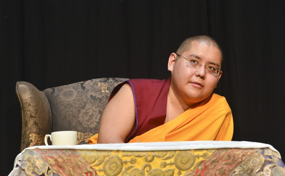 His Eminence Ling Rinpoche seated on stage leaning to his left and half smiling in thought.