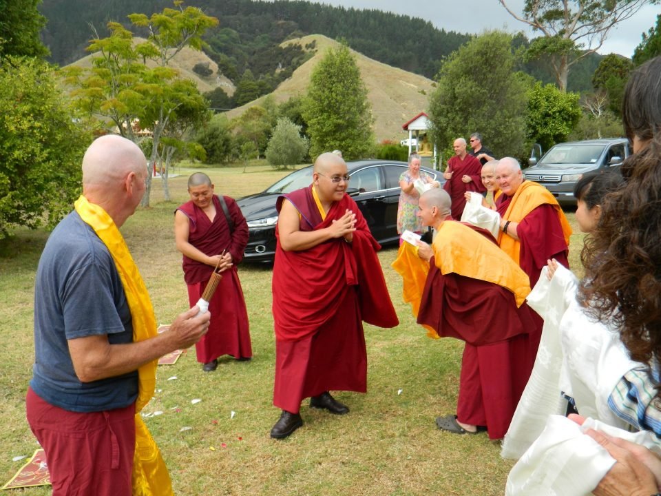 His Eminence Ling Rinpoche walking from a parked car across a lawn with hands folded in greeting being welcomed by a small group of western nuns and lay people holding khatas and smiling.