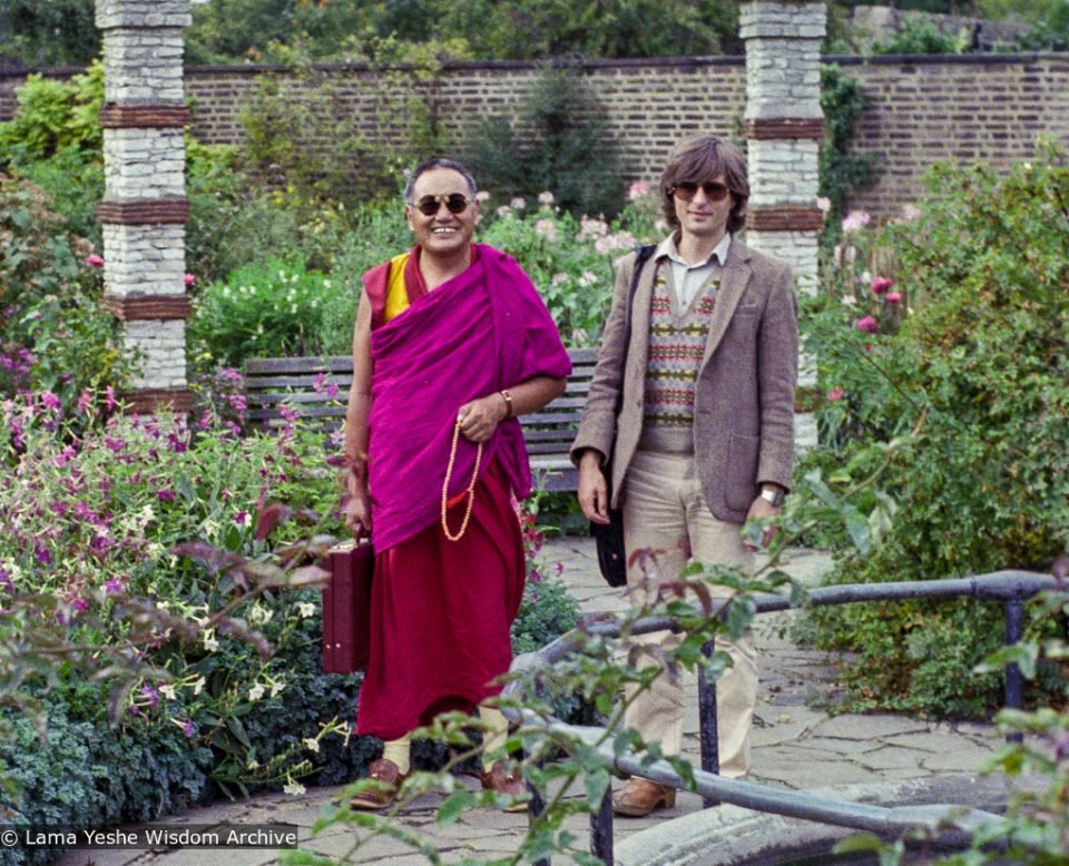 Lama Yeshe in monks robes standing next to Geoff Jukes in sunglasses and a tweed sport coat in an English garden