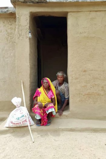 Two people seated inside of the doorway of a mud walled home with a long white stick and large bag nearby.