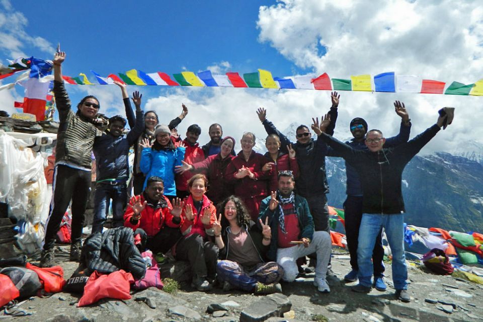 Tushita team and friends from Spain standing together on the top of Drilbu Ri mountain with hands raised above their heads smiling and cheering with prayer flags flapping overhead on a bright sunny day.