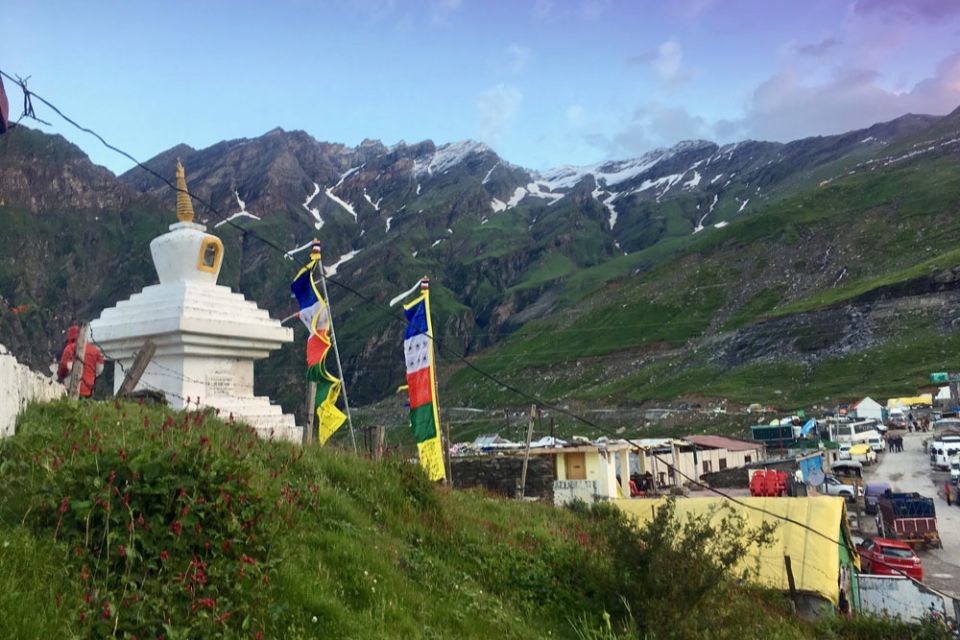 White stupa surrounded by poles of prayer flags in a small village with mountains in the background.