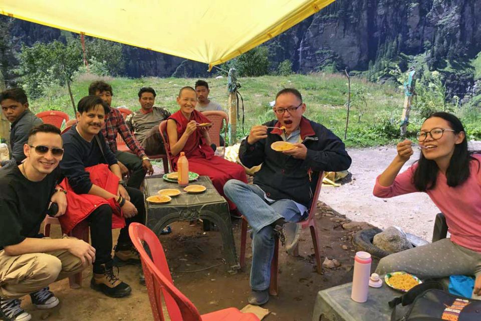 Tushita staff smiling while seated on red plastic chairs underneath a tent on the roadside eating food from plates held in hands and on makeshift tables.