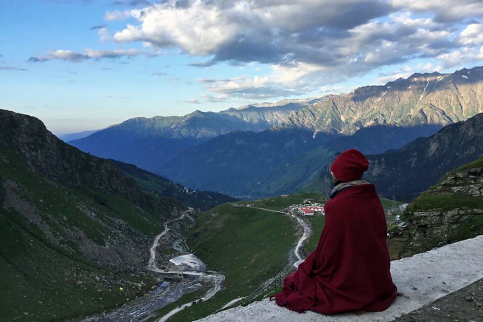 Person dressed in a winter hat with a shawl wrapped around them seated on a ledge overlooking a valley with rivers and mountains all around and clouds in a blue sky.