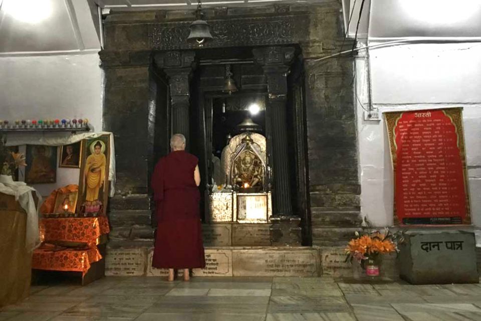 A monastic standing in front of the Chenrezig statue with hands folded together in prayer inside of a white walled Phakpa temple.