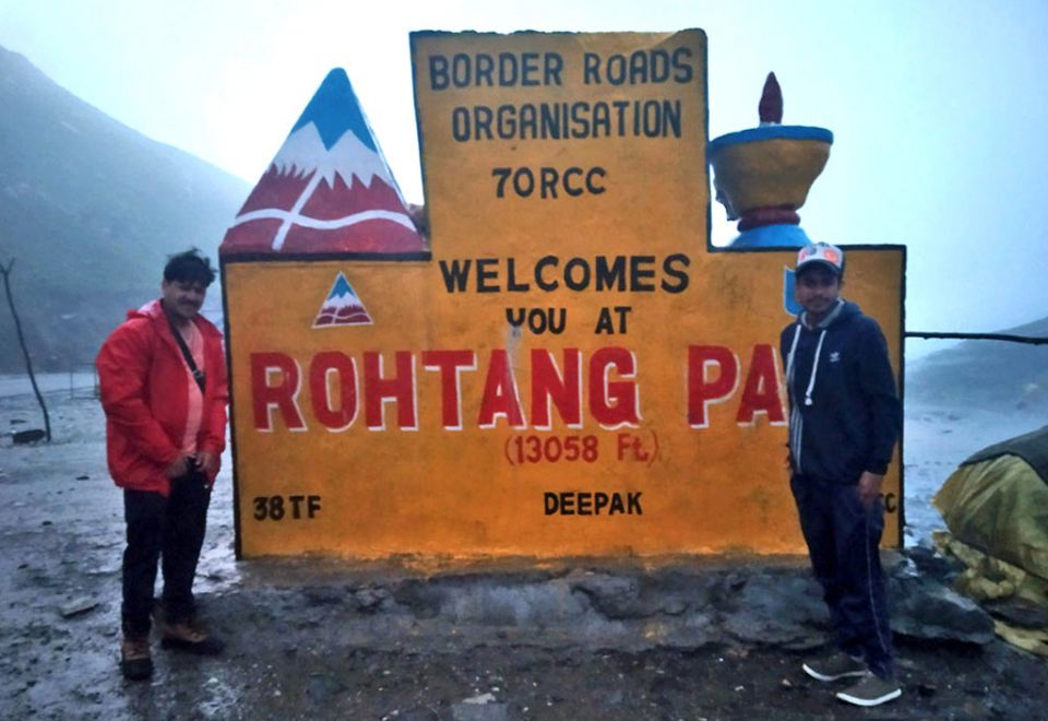 Two people standing on either side of a very large yellow concrete sign that says welcome to rohtang pass underneath an overcast sky.