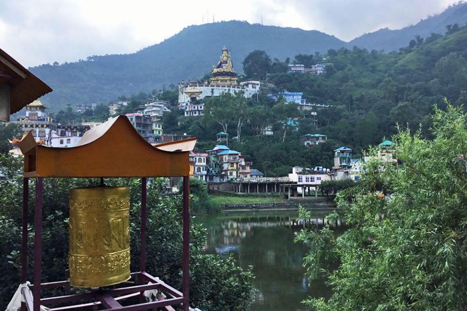 A large golden colored prayer wheel in the foreground and the lake in the midground and the gigantic Padmasambhava statue in background with mountains in the backdrop.