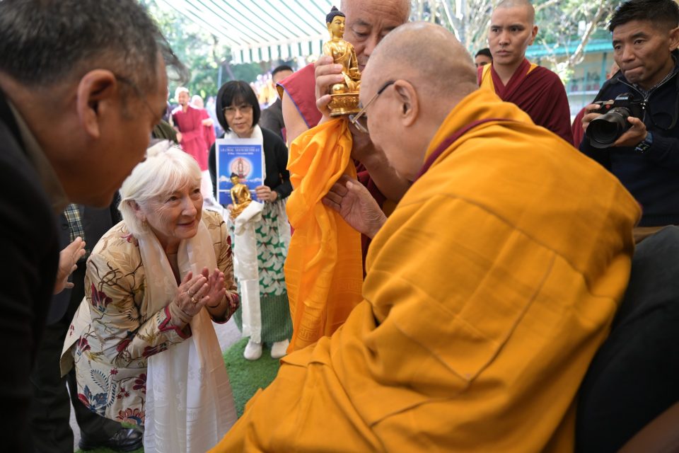 A smiling woman with palms joined bows toward the Dalai Lama as a monk presents a Buddha statue.