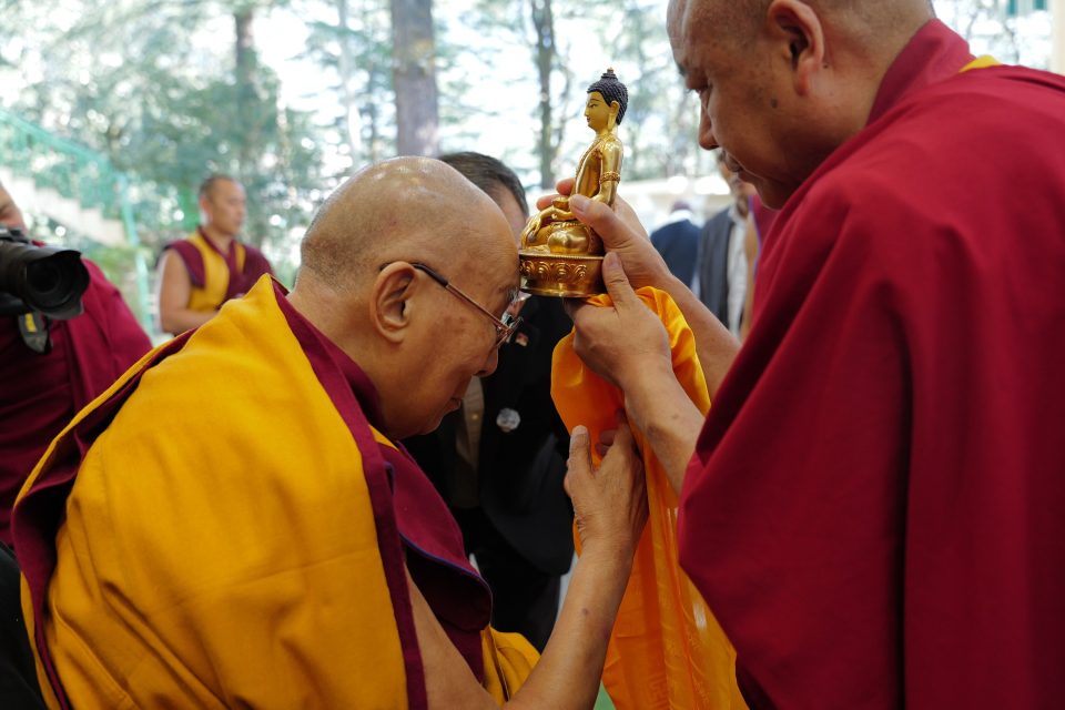 The Dalai Lama bows his head to touch a golden Buddha statue held out to him by a monk.
