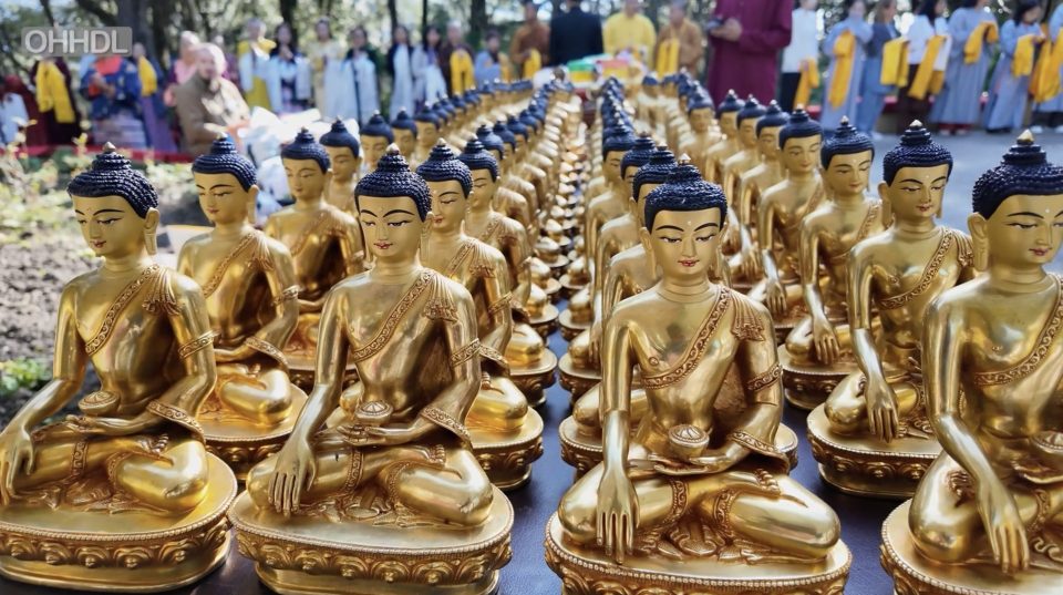 Rows of gold Buddha statues with dark blue hair, with a crowd holding ceremonial scarves behind them.